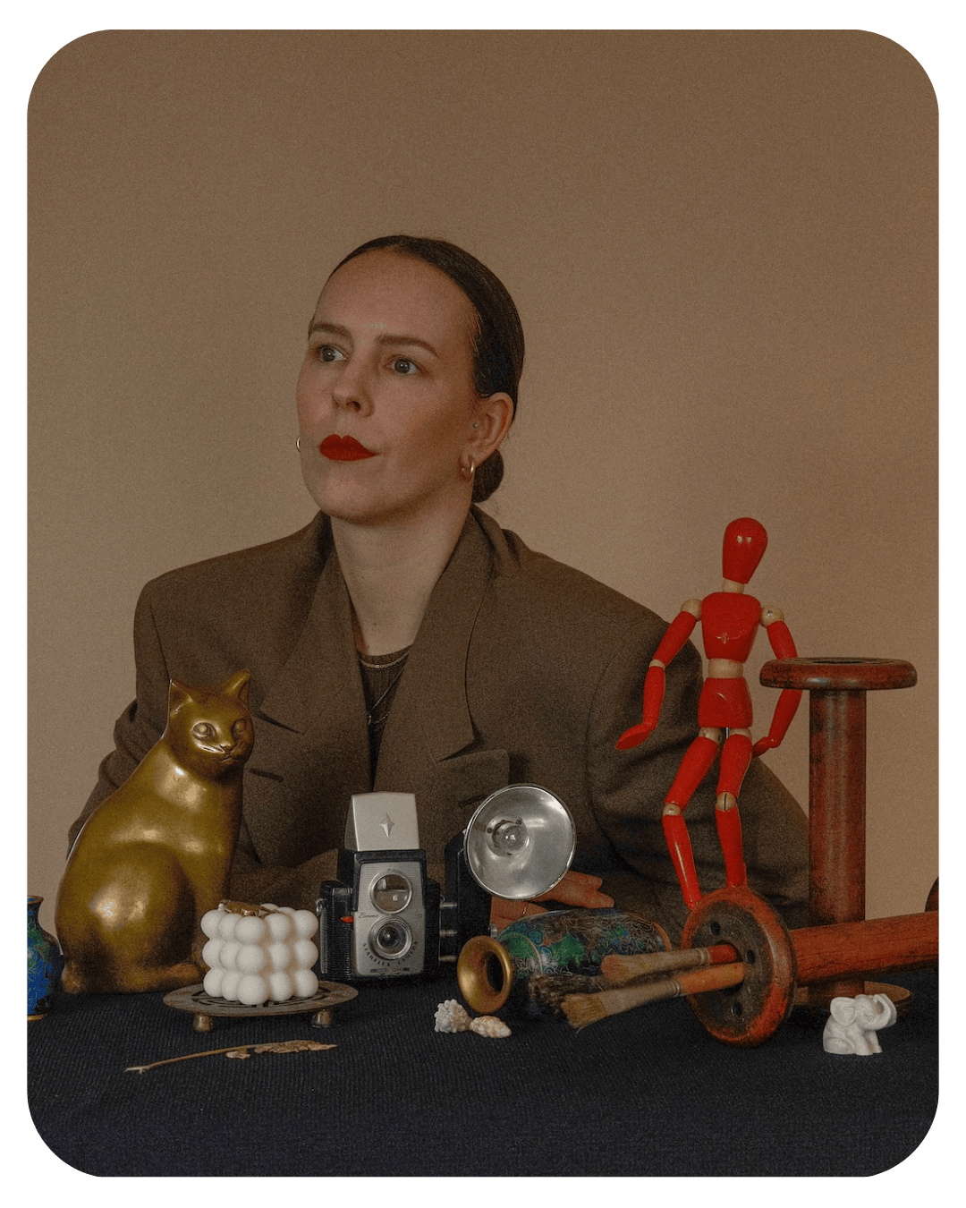 Woman in a brown blazer surrounded by vintage objects like a camera, red mannequin, and golden cat statue.