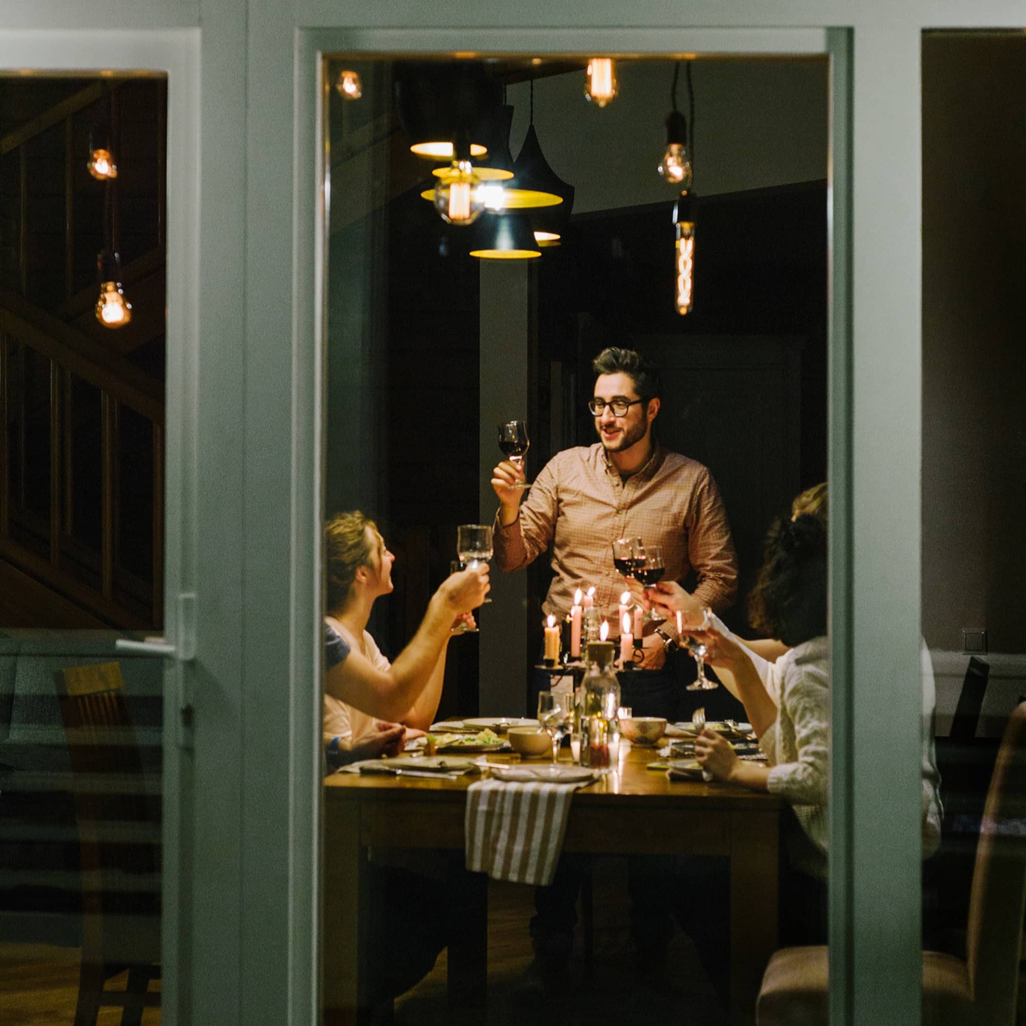 Man standing to give a toast with wine glasses at a warm candlelit dinner party with friends seen through a window.