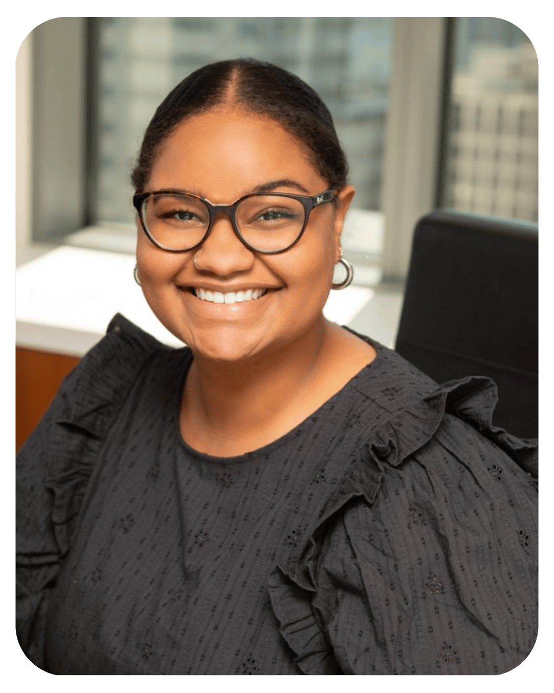 Smiling professional woman with glasses wearing a black ruffled blouse in an office setting with city view.