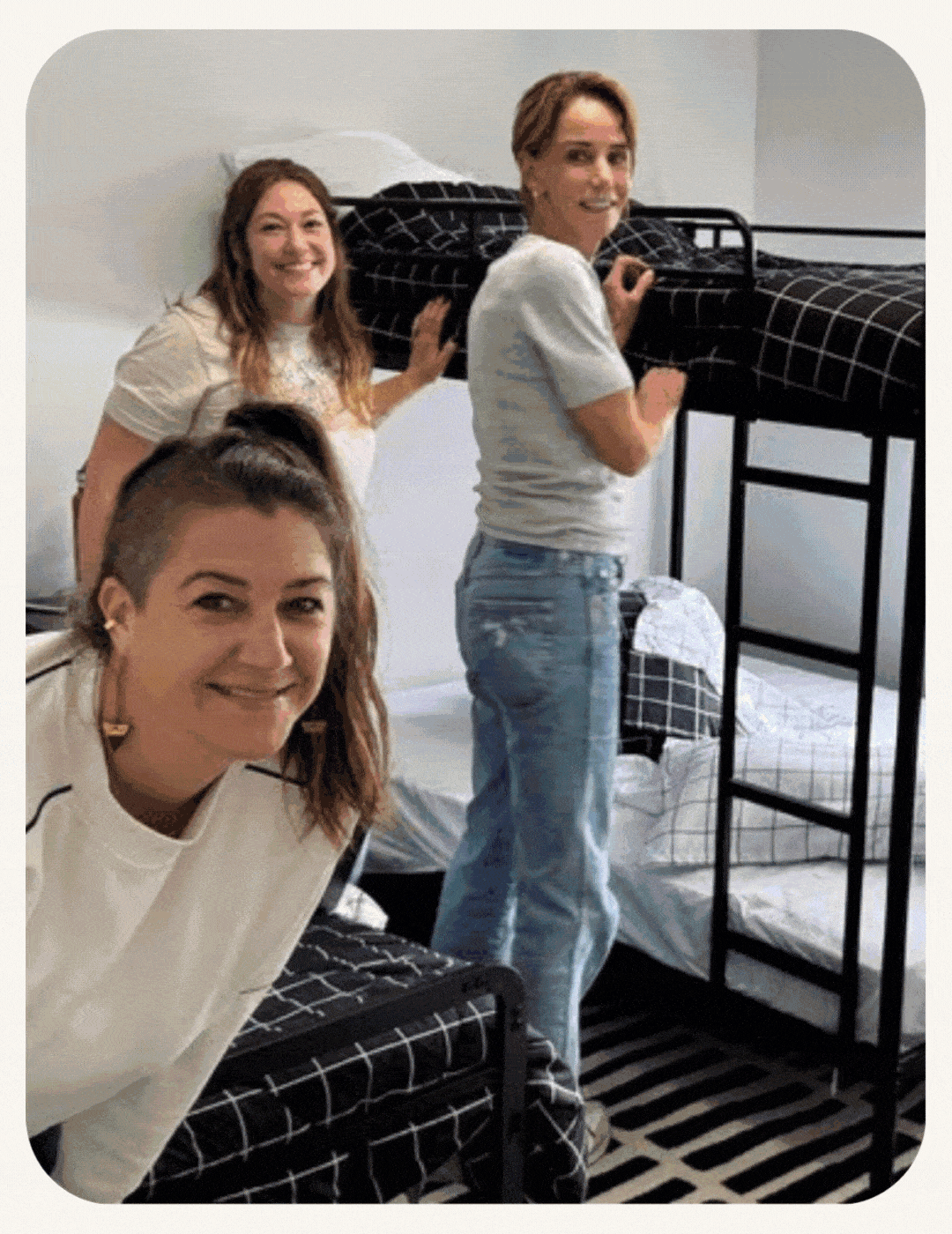 Three women smiling while setting up black metal bunk beds with grid-patterned bedding in a bedroom.